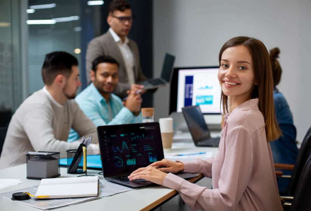 A smiling businesswoman works on her laptop while her colleagues discuss business in the background.