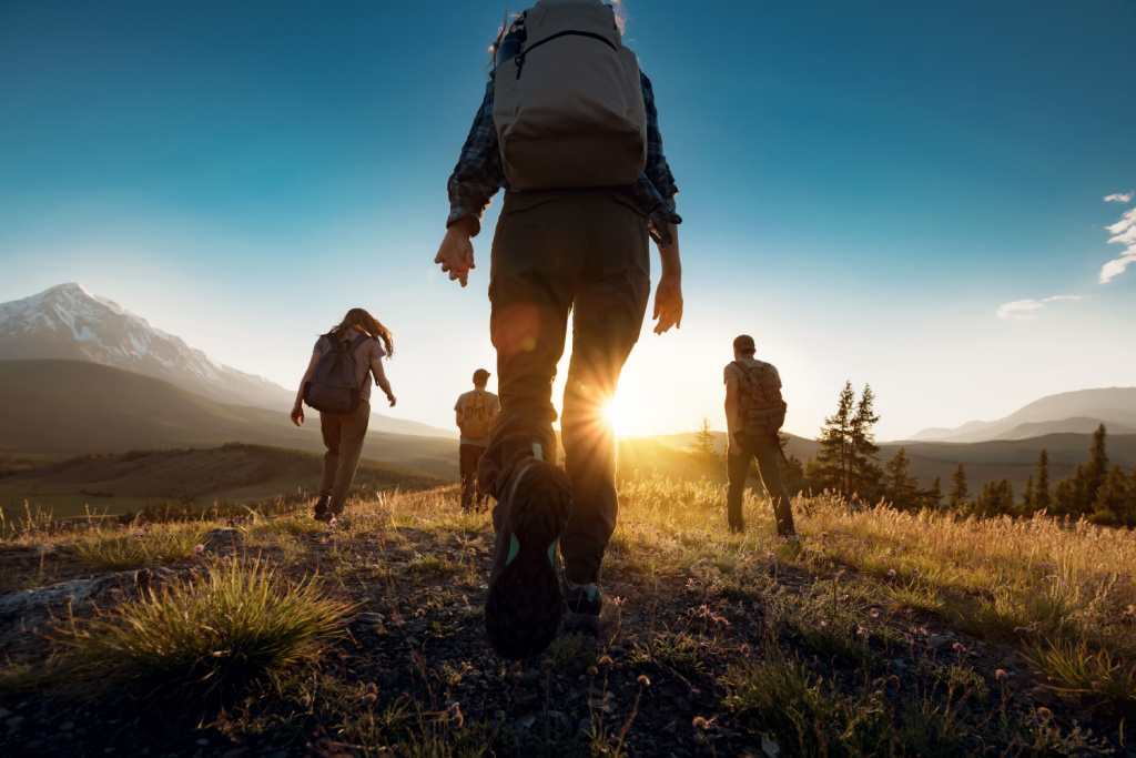 Four people with backpacks hike up a hill at sunset with a mountain range in the distance.