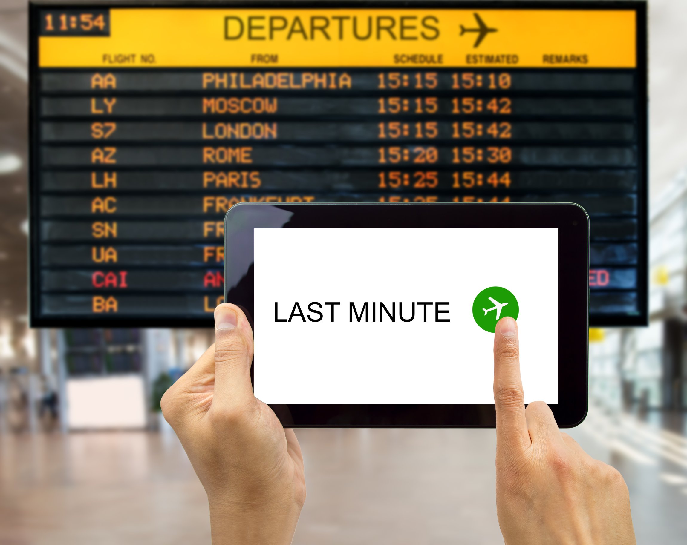 A hand holding a tablet with the text 'Last Minute' on the screen in front of an airport departures board.