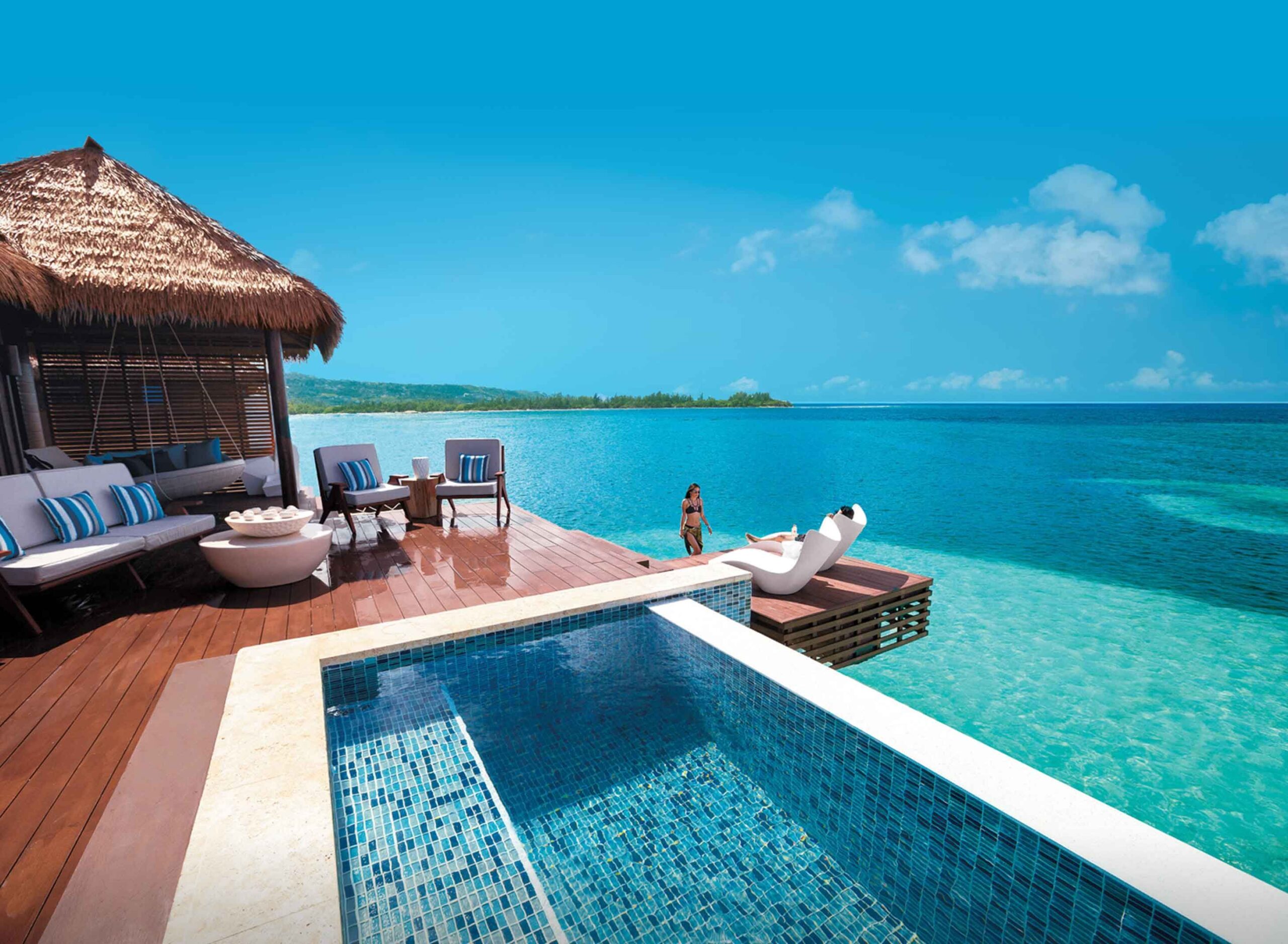 A woman in a bikini is relaxing on a lounger on a wooden dock that extends out into the blue ocean. There is a thatch-roofed cabana on the dock and a plunge pool built into the deck. The water is crystal clear and there is a blue sky with a few white clouds in the background. The image represents the search query 'Luxury vacation packages offering exclusive accommodations and amenities tailored to discerning travelers'.