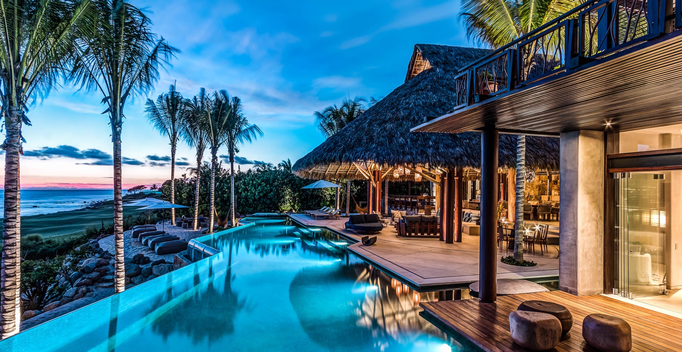 A luxury villa with an infinity pool overlooking the ocean at sunset with palm trees and a thatched roof, representing the search query 'Luxury travel destinations and experiences for 2024'.