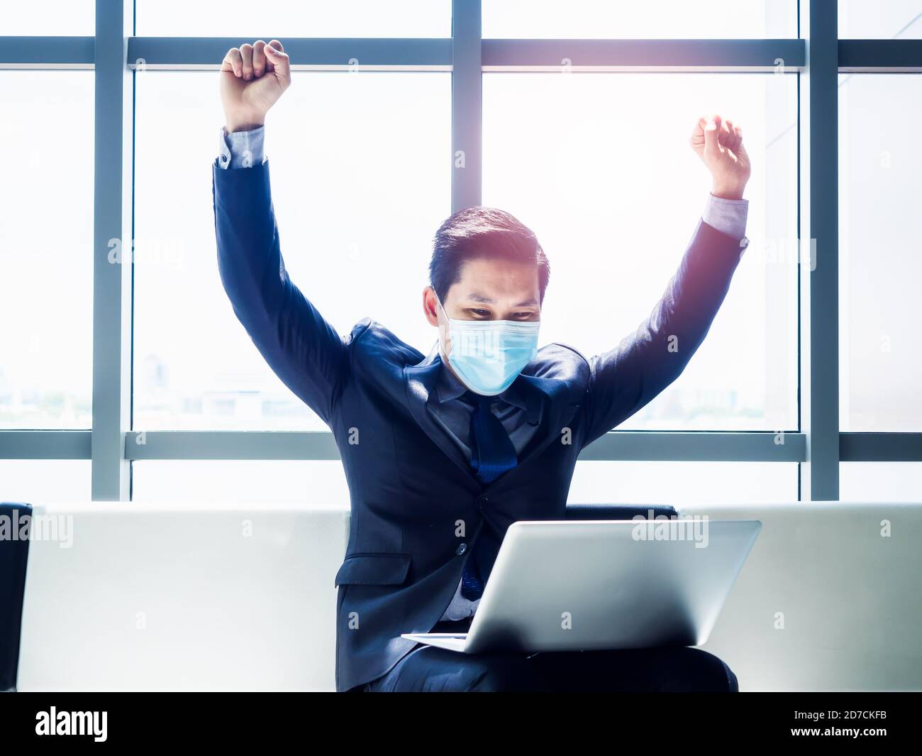 A businessman wearing a suit and a medical mask celebrates while sitting in front of an open laptop with a padlock icon on the screen.