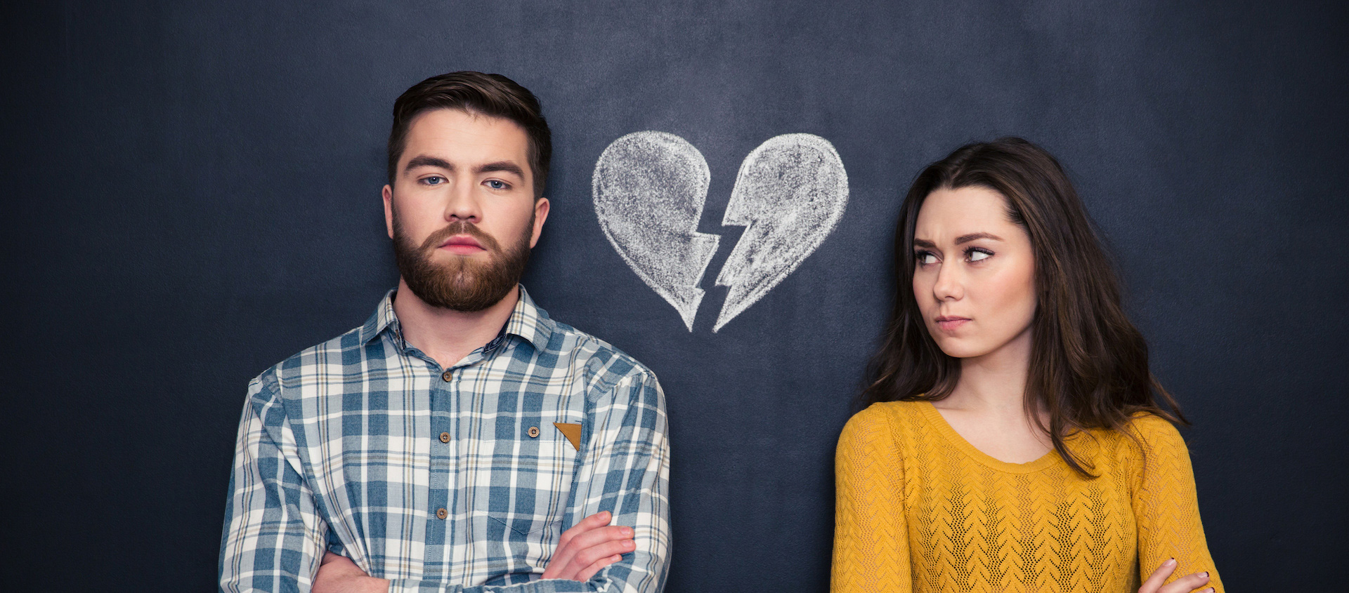 A man and a woman stand in front of a chalkboard with a broken heart drawn on it; they both have their arms crossed and are looking away from each other, representing the emotional toll of divorce.