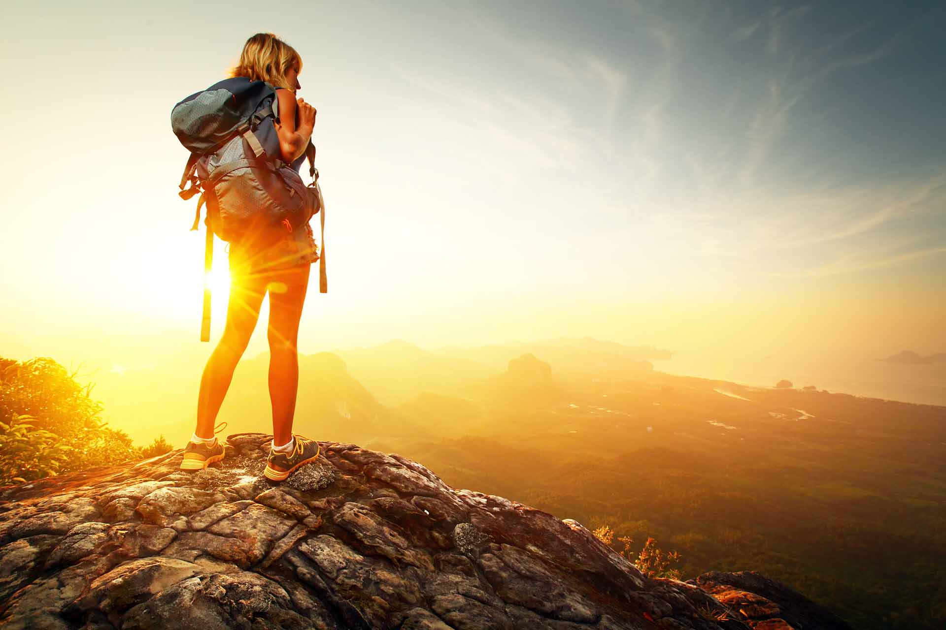 A woman standing on a rock and looking at the sunrise with a large backpack on her back.