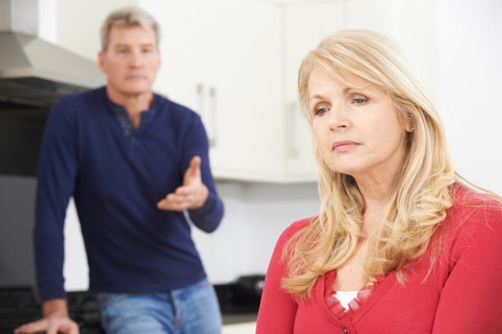 A blonde woman in a red shirt looks sadly away from the camera while a man in a blue shirt stands in the background.