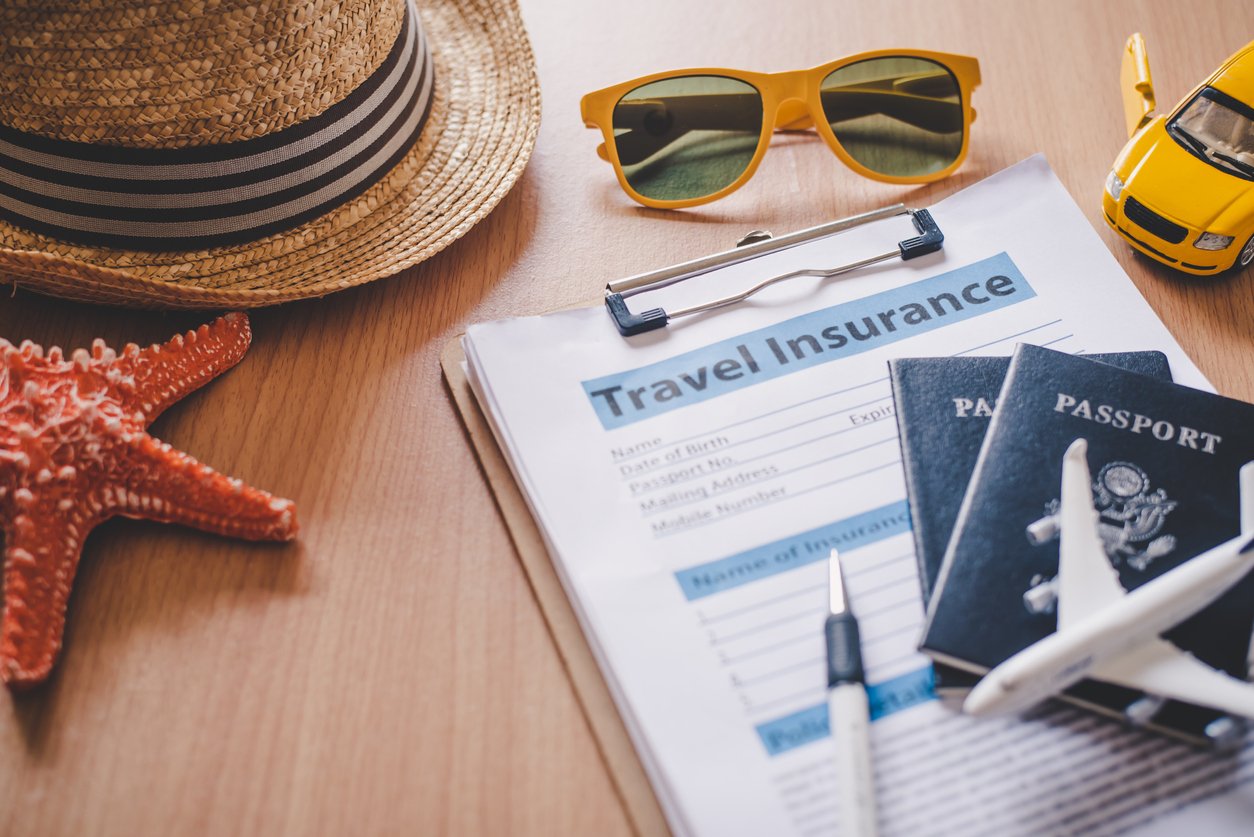 A straw hat, sunglasses, toy car, passport, plane, and travel insurance form on a table.